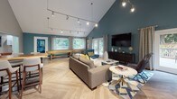 This thoughtfully designed single-floor gathering space combines modern aesthetics with functional amenities, making it an inviting destination for socializing and recreation. The main lounge area immediately impresses with its dramatic vaulted ceilings painted in a rich teal, contrasted by crisp white rafters and a sleek track lighting system that illuminates the room from above. Light-toned herringbone-patterned flooring flows throughout, tying the entire space together cohesively.

The lounge area is furnished with a plush sofa, accent chairs in coordinating teal upholstery, a round side table, and a large wall-mounted flat-screen television atop a dark media console. A full-length shuffleboard table anchors the game area, adding an element of entertainment. French doors and large windows along the perimeter flood the interior with natural light and offer views of an adjacent outdoor patio and pool area.

A dedicated dining nook accommodates a dark-topped table with woven rattan chairs, surrounded by windows that frame lush outdoor scenery. The kitchenette is well-appointed with dark cabinetry, white quartz countertops, a stainless steel sink, a dishwasher, a coffee station, and an elegant stone-tile backsplash with open floating shelves for display. Together, these spaces create a warm, versatile, and visually striking communal environment.