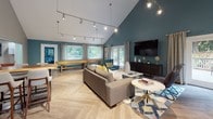 This thoughtfully designed single-floor gathering space combines modern aesthetics with functional amenities, making it an inviting destination for socializing and recreation. The main lounge area immediately impresses with its dramatic vaulted ceilings painted in a rich teal, contrasted by crisp white rafters and a sleek track lighting system that illuminates the room from above. Light-toned herringbone-patterned flooring flows throughout, tying the entire space together cohesively.

The lounge area is furnished with a plush sofa, accent chairs in coordinating teal upholstery, a round side table, and a large wall-mounted flat-screen television atop a dark media console. A full-length shuffleboard table anchors the game area, adding an element of entertainment. French doors and large windows along the perimeter flood the interior with natural light and offer views of an adjacent outdoor patio and pool area.

A dedicated dining nook accommodates a dark-topped table with woven rattan chairs, surrounded by windows that frame lush outdoor scenery. The kitchenette is well-appointed with dark cabinetry, white quartz countertops, a stainless steel sink, a dishwasher, a coffee station, and an elegant stone-tile backsplash with open floating shelves for display. Together, these spaces create a warm, versatile, and visually striking communal environment.