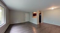 This single-floor unit is designed with a practical, open layout that balances living and functional spaces effectively. The main living area is generously proportioned, finished with dark hardwood-style flooring that runs throughout, lending a warm, polished aesthetic. Neutral gray walls complement the flooring and create a clean, versatile backdrop. A front entry door opens directly into this open space, which connects seamlessly to the kitchen via a pass-through window framed by a partial wall — a design detail that maintains an open feel while defining each area.

The kitchen is well-equipped with rich dark cabinetry, marble-style countertops, a white tile backsplash, a black gas range, and a black refrigerator. Hardwood-style flooring continues into this space, maintaining visual continuity. A separate bedroom features plush carpeting and a wide window overlooking lush greenery, offering abundant natural light and pleasant outdoor views. Baseboard heating units line the walls in both the bedroom and the expansive windowed living area.

The bathroom is finished with light marble-style wall tile surrounding a full soaking tub, complemented by a white vanity with under-cabinet storage and a mirrored medicine cabinet, creating a bright, clean environment throughout.