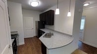 This single-floor residence offers a thoughtfully designed layout with modern finishes throughout. The kitchen serves as a central highlight, showcasing rich dark cabinetry that provides ample storage and a striking contrast against the speckled granite countertops. A curved peninsula extends into the living area, creating a natural division of space while offering additional workspace and a casual dining option. Pendant lights hang above the peninsula, casting a warm, focused glow that enhances the kitchen's contemporary aesthetic. A stainless steel refrigerator and integrated appliances complete the functional cooking space, while a single-basin undermount sink adds a clean, polished look. The kitchen flows seamlessly into the adjoining areas, with durable wood-style flooring running throughout and lending warmth to the interior. Neutral-toned walls keep the space feeling bright and versatile. A white paneled door with an arched detail provides access to an adjacent room or storage area, adding a classic architectural touch. The overall ambiance is clean, modern, and move-in ready, balancing practical functionality with contemporary design sensibilities suited for everyday living.
