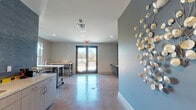 This single-floor space is designed with a clean, contemporary aesthetic that blends functionality with thoughtful design details. Polished concrete floors run throughout, offering a sleek, durable surface that reflects natural light and adds an industrial-modern character to the interior.

The main open area is defined by a striking blue accent wall adorned with a sculptural wall art piece, creating a bold visual focal point. Opposite this wall, a textured blue elongated-hexagon tile feature wall houses a wall-mounted flat-screen display above a built-in floating counter with bar stool seating — ideal for collaborative work or casual use.

Large glass double doors open directly to the outdoors, flooding the space with abundant natural light and providing clear views of the surrounding exterior. A generous window in an adjacent area further enhances the bright, airy atmosphere and frames views of the landscaped grounds outside.

The space also includes a well-appointed utility area featuring light wood cabinetry, stone-look countertops with a deep stainless steel sink, and a full blue tile backsplash that mirrors the design language seen throughout. A mobile workbench with a butcher block top adds flexible, functional workspace. Recessed lighting supplements the natural light, ensuring the interior remains well-lit throughout. Overall, the space conveys a polished yet practical environment suited for a variety of uses.