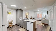 This thoughtfully designed two-bedroom, one-bathroom residence offers a functional and contemporary layout across a single floor. The open-plan living and kitchen area serves as the heart of the home, featuring modern flat-front cabinetry in a warm grey-wood finish, white countertops, a built-in dishwasher, and a freestanding range. A peninsula with a sink provides additional workspace and connects the kitchen seamlessly to the living area, which receives natural light through a window overlooking an exterior courtyard. Wood-style plank flooring runs throughout the entire space, adding warmth and visual continuity. The primary bedroom is a comfortable retreat, featuring a large window with vertical blinds, clean white walls, and ample room for a full-sized bed. The second bedroom is versatile, accommodating bunk beds alongside a desk area, making it well-suited as a shared or multifunctional room. Both bedrooms benefit from natural light. The bathroom is clean and practical, offering a full soaking tub with a shower, open shelving, and a striped curtain for privacy. A dedicated walk-in closet with full-length hanging rods provides generous storage. The overall design aesthetic is modern and understated, with a neutral palette and efficient use of space throughout.