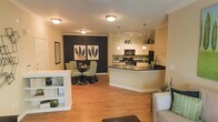 This single-floor residence showcases a thoughtfully designed open-concept layout that connects the living, dining, and kitchen areas into one cohesive, flowing space. Warm hardwood flooring runs throughout, lending a sense of continuity and natural warmth to the interior.

The kitchen is a functional centerpiece, featuring dark countertops that contrast elegantly with cream-toned cabinetry. A peninsula with a built-in sink provides additional prep space and a natural gathering point. Black appliances, including a microwave and refrigerator, complement the bold color palette, while pendant lighting above the peninsula adds a refined, modern touch.

Adjacent to the kitchen, the dining area is defined by a striking dark accent wall that serves as a bold focal point. A round dining table surrounded by sage green upholstered chairs creates an intimate yet stylish setting, anchored by a circular area rug.

The living area benefits from abundant overhead lighting and crown molding details that lend a polished, finished aesthetic. A white open shelving unit provides a subtle visual divider between spaces while offering display and storage options.

Overall, the space strikes a balance between modern design and comfortable functionality, with thoughtful lighting, a cohesive neutral color scheme, and a layout that encourages easy movement and interaction throughout.