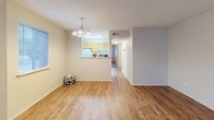 This single-floor residential unit offers a practical and well-organized layout across several distinct living areas. The open living and dining area features warm wood-style laminate flooring and a chandelier light fixture overhead, creating a welcoming atmosphere. A half-wall pass-through connects this space to the kitchen, which is equipped with light maple cabinetry, ample upper and lower storage, a coil-top electric range, a full-size refrigerator, and a built-in range hood. A dedicated dining area adjacent to the kitchen benefits from natural light through a window with outdoor views.

The bedrooms are finished with neutral-toned carpet and feature sliding-door closets for storage. Windows allow natural light to filter in, and the textured ceilings are consistent throughout the unit. The bathroom includes large-format light gray tile surrounding a full soaking tub and shower combination, a wall-mounted chrome towel rack, a toilet, and a vanity with a countertop sink. A hallway connects the primary living spaces to the private rooms, contributing to a logical and efficient floor plan overall.