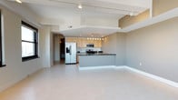 This contemporary two-floor space is thoughtfully designed with an open-concept layout that promotes a seamless flow between living and kitchen areas. The kitchen is a standout feature, outfitted with light natural wood cabinetry, dark countertops, and a full suite of stainless steel appliances including a side-by-side refrigerator with ice and water dispenser, a microwave, and a gas range. A central kitchen island with a built-in sink provides additional prep space and serves as a natural dividing element between the kitchen and the expansive open living area. The floors throughout are covered in large-format light-toned tiles that lend a clean, polished aesthetic. Recessed lighting and track lighting mounted to the ceiling ensure the space is well-illuminated, while structural ceiling details add architectural character. The overall color palette is neutral and refined, with soft white and warm gray tones creating a calm and inviting ambiance. A hallway visible from the main area suggests additional rooms and provides access to the rest of the two-floor layout, offering ample living space for a variety of uses.