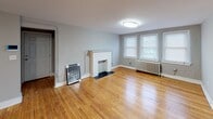 This single-floor unit blends classic architectural character with functional updates throughout. The living room serves as a welcoming centerpiece, highlighted by warm, honey-toned hardwood floors, a charming white-mantled fireplace, and multiple windows that invite natural light into the space. A cast-iron radiator adds a nod to the building's heritage while providing reliable warmth. The adjacent bedroom echoes the same hardwood flooring and features its own cast-iron radiator and double windows fitted with blinds, creating a bright yet private retreat. The kitchen is equipped with dark espresso-toned cabinetry, light countertops, and stainless steel appliances including a gas range and an over-the-range microwave. Wood-look flooring completes the kitchen's clean, practical aesthetic. The bathroom is a standout space, finished with large-format marble-patterned tile covering both the floors and the tub surround. A soaking tub, a compact vanity, and a window above the tub round out this well-appointed room. Throughout the unit, consistent neutral gray walls tie each space together, creating a cohesive and move-in-ready environment that balances timeless details with modern conveniences.