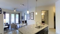 This single-floor residence showcases an open-concept layout that integrates the kitchen, dining, and living areas into one cohesive, flowing space. The kitchen is anchored by a substantial granite-topped island with a stainless steel undermount sink, complemented by dark cabinetry that provides a striking contrast to the light stone countertops. Overhead, a combination of track lighting and elegant pendant lights illuminate the workspace effectively.

The open layout transitions naturally into the dining area, where a round table and seating arrangement sit beneath a ceiling fan, adding both function and comfort. Large windows dressed with blinds and draping curtains allow generous natural light to fill the space, creating a bright and welcoming ambiance throughout.

Neutral white walls adorned with framed artwork contribute a clean, contemporary aesthetic, while tile and carpet flooring delineate the different functional zones. A glimpse into an adjacent room reveals additional living or bathroom space, suggesting a well-organized and thoughtfully designed floor plan. The overall atmosphere is modern, airy, and functionally arranged.