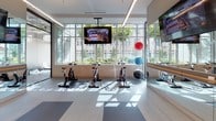 Located at 825 S Hill St in the heart of Downtown Los Angeles, this contemporary fitness center offers a comprehensive, single-floor layout designed to support a full range of workout disciplines. The facility is organized into distinct training zones, each well-appointed and thoughtfully arranged. The strength training area features an extensive dumbbell rack, multiple flat benches, and a row of power racks and cable machines, all set against floor-to-ceiling mirrors and motivational wall graphics. The free weights zone is complemented by large windows that flood the space with natural light and frame views of the surrounding urban landscape. A dedicated cardio section houses treadmills, ellipticals, rowing machines, and stationary bikes, positioned along expansive window walls to maximize daylight exposure. A separate cycling studio offers spin bikes, stability balls, and wall-mounted TVs, with lush outdoor greenery visible through the windows. Throughout the facility, exposed ceilings with industrial-style ductwork and linear LED lighting contribute to a clean, high-energy aesthetic. The light wood-toned flooring and neutral grey palette create a bright, motivating environment. Multiple flat-screen TVs are mounted throughout to keep members engaged during their workouts.