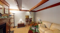 This well-appointed 1-bedroom, 1-bathroom unit is thoughtfully laid out across a single floor, offering a comfortable and inviting living experience. The living room serves as the heart of the space, featuring rich wood-stained exposed ceiling beams set against a textured ceiling, adding a rustic yet refined character. A wood-paneled wall frames a classic fireplace flanked by built-in shelving, creating a warm focal point for the room. Carpeted floors and neutral-toned walls contribute to the cozy ambiance, while a set of French doors allows natural light to filter in and provides direct access to an outdoor area. Adjacent to the living room, a bar-height dining nook with wooden bar stools offers a casual dining option. The bedroom is a quiet retreat, featuring a louvered wood closet, a window dressed with decorative curtains, and ample space for a full-sized bed. The full bathroom is clean and functional, complete with a single-sink vanity with wood cabinetry, a wide mirror with overhead lighting, and a full tub-shower combination. This unit blends warm wood accents, soft neutral tones, and practical design elements to create a comfortable and characterful living space.