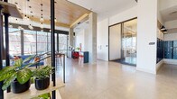 This thoughtfully designed single-floor commercial space blends industrial and contemporary elements to create a functional and visually engaging environment. Polished concrete floors run throughout, reflecting natural light and lending a clean, modern aesthetic to the open layout. Warm wood-paneled ceiling sections contrast beautifully with crisp white walls and exposed concrete columns, creating a layered architectural character.

The main open area serves as a central hub, featuring a bar-height counter with bold red seating and a dedicated coffee station with sleek gray cabinetry. Floor-to-ceiling windows along the perimeter provide abundant natural light and expansive outdoor views, while a striking cluster of Edison-style pendant lights anchors the social area beneath the wood ceiling. A suspended cylindrical fireplace adds a distinctive focal point to the lounge zone, which is furnished with comfortable seating.

A glazed private office or meeting room with sliding glass doors provides enclosed workspace without sacrificing visual openness. A comfortable lounge nook near large windows offers a quiet retreat with leather seating and indoor greenery. The space also includes elevator access and a stairwell entry, supporting seamless circulation throughout.