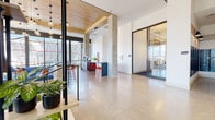 This thoughtfully designed single-floor commercial space blends industrial and contemporary elements to create a functional and visually engaging environment. Polished concrete floors run throughout, reflecting natural light and lending a clean, modern aesthetic to the open layout. Warm wood-paneled ceiling sections contrast beautifully with crisp white walls and exposed concrete columns, creating a layered architectural character.

The main open area serves as a central hub, featuring a bar-height counter with bold red seating and a dedicated coffee station with sleek gray cabinetry. Floor-to-ceiling windows along the perimeter provide abundant natural light and expansive outdoor views, while a striking cluster of Edison-style pendant lights anchors the social area beneath the wood ceiling. A suspended cylindrical fireplace adds a distinctive focal point to the lounge zone, which is furnished with comfortable seating.

A glazed private office or meeting room with sliding glass doors provides enclosed workspace without sacrificing visual openness. A comfortable lounge nook near large windows offers a quiet retreat with leather seating and indoor greenery. The space also includes elevator access and a stairwell entry, supporting seamless circulation throughout.