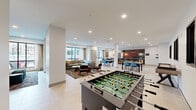 Located at 260 Portland Ave in downtown Minneapolis, this contemporary community lounge is a well-appointed, single-floor amenity space designed for social interaction and relaxation. The open-plan layout is anchored by clean white walls, light-toned large-format tile flooring, and recessed lighting throughout, lending the space a bright and modern character.

The living area offers a comfortable sectional sofa and cushioned armchairs arranged around a coffee table, complemented by a wall-mounted flat-screen TV. Large floor-to-ceiling windows frame urban views of the Minneapolis skyline, allowing generous natural light to fill the room. Sheer drapery softens the look while maintaining the open ambiance.

A dedicated lounge nook features a built-in banquette with channel-tufted upholstery, dark accent tables, and a textured tile feature wall, creating an intimate setting within the broader open space. Pendant lighting adds a warm, stylish touch above the central island and dining tables, which are surrounded by bold blue chairs, offering ample seating for group gatherings.

The game area is well-equipped with a foosball table and a shuffleboard table, providing recreational options for residents. The overall design balances industrial-modern elements with inviting furnishings, making this a versatile and engaging communal space.
