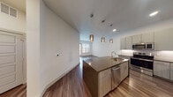 This single-floor unit showcases a contemporary design with an open-concept layout that seamlessly connects the kitchen, dining, and living areas. The kitchen is a standout feature, equipped with two-tone cabinetry in soft gray and warm wood tones, dark quartz countertops, and a full suite of stainless steel appliances including a French door refrigerator, electric range, built-in microwave, and dishwasher. A large central island with an undermount sink serves as the focal point, topped with matching quartz and illuminated by a trio of brushed gold cylindrical pendant lights. A white subway tile backsplash and under-cabinet lighting add refined detail. Rich wood-look flooring extends throughout the main living areas, anchoring the space with warmth. The living area benefits from natural light through a large window and sliding glass door, complemented by a ceiling fan. The bedroom features plush carpet, a ceiling fan, and generous closet space. The bathroom is well-appointed with a double-sink vanity topped in granite, wood-tone cabinetry, a soaking tub with tile surround, and a separate glass-enclosed shower, all finished with wood-look flooring for a cohesive, polished aesthetic.