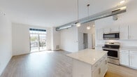 This bright and contemporary single-floor unit showcases a thoughtfully designed open-concept layout that seamlessly connects the kitchen, dining, and living areas. Light-toned hardwood flooring runs throughout, complementing the clean white walls and creating a cohesive, airy aesthetic.

The kitchen is a standout feature, offering white shaker-style cabinetry with brushed gold hardware, marbled quartz countertops extending to a generous island with pendant lighting overhead, and a full suite of stainless steel appliances including a range, microwave, dishwasher, and refrigerator. An exposed industrial-style duct adds a loft-inspired character to the space.

The living area is filled with natural light from large windows and a glass-paneled balcony door that opens to an outdoor area with views of the surrounding neighborhood. The bedrooms are well-proportioned, each benefiting from natural light through sizeable windows framing pleasant exterior views.

The unit includes two bathrooms — one featuring a walk-in shower, white vanity with quartz countertop, and wood-look flooring; the second is a spacious, accessible bathroom with a wide vanity and generous floor space. Clean white finishes throughout both bathrooms maintain the unit's consistent modern design language.