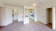 This single-floor unit offers a practical and well-organized layout across several rooms. The living area is carpeted and open, connecting directly to the kitchen via a half-wall peninsula that provides a pass-through counter with a sink. The kitchen is equipped with white appliances, including a refrigerator, microwave, and electric range, alongside cabinetry with wood-toned accents and tile flooring. A sliding glass door fitted with vertical blinds in the living area allows natural light to enter and provides outdoor access. The bedrooms are carpeted and feature neutral walls, each with windows fitted with blinds that allow natural light while maintaining privacy. The unit includes two bathrooms: one features a standalone walk-in shower with a frosted glass door and chrome frame, accompanied by a vanity and toilet; the second bathroom offers a full shower-tub combo with a curtain rod, vanity with mirror, and toilet. Throughout the unit, the color palette remains consistent with soft beige and white tones, contributing to a clean, understated aesthetic. The single-floor design ensures ease of navigation throughout the space.