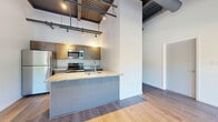 Located in Rochester's vibrant East End neighborhood, this one-bedroom, one-bathroom unit at 50 Prince Street showcases a striking industrial-loft aesthetic. Soaring ceilings with exposed steel beams, corrugated metal details, and track lighting define the open living and kitchen area, creating a bold, contemporary atmosphere throughout.

The kitchen is well-appointed with stainless steel appliances — including a refrigerator, electric range, and built-in microwave — complemented by warm taupe cabinetry and light granite countertops. A peninsula with a stainless steel sink adds functional prep space and serves as a natural divider between the kitchen and the combined dining and living area. Wood-look flooring flows seamlessly throughout the unit.

The open living and dining space is bright and airy, featuring oversized windows with roller shades that flood the room with natural light and frame views of the surrounding Rochester streetscape and mature trees. The bedroom offers a comfortable footprint with a sliding-door closet and additional access to multiple closets for generous storage.

The bathroom is clean and minimalist, featuring wood-look flooring, crisp white walls, and simple fixtures. Two hallways connect the spaces efficiently. This unit offers a well-balanced blend of industrial character and modern finishes in a prime urban Rochester location.
