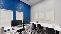 Located at 400 Putnam Dr in Tallahassee, FL, this well-appointed business center is designed to support both focused individual work and collaborative group activity. The space is defined by a striking cobalt blue 3D wave-textured accent wall that serves as a bold focal point, contrasting dynamically with the soft gray tones of the surrounding walls and light wood-look flooring. Two white workstations are positioned along the accent wall, each equipped with dual monitors on articulating mounts, providing a fully functional computing environment for residents or guests. Natural light filters in through windows fitted with white horizontal blinds, keeping the atmosphere bright and productive. A central conference area features a clean white table surrounded by modern black chairs, anchored by a bold black-and-white geometric area rug. A large wall-mounted flat-screen display makes this zone ideal for presentations or collaborative viewing. Decorative framed sun-burst artwork adds a warm, curated aesthetic to the neutral walls. A prominent gold Roman numeral clock adorns one wall, adding a sophisticated design touch. The space is accessed through a glass-paneled interior door, maintaining visual connectivity while offering a defined, purposeful environment for work and meetings.