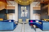 Located on Delmar Boulevard in University City, Missouri, this striking hospitality space delivers a bold and energetic design experience across a single expansive floor. The grand lobby immediately commands attention with soaring double-height ceilings, floor-to-ceiling windows that flood the space with natural light, and a dramatic cascading gold chandelier. A signature "PLAY" wall installation serves as the focal point, backed by a deep blue feature wall and flanked by plush royal blue velvet sofas with vibrant accent pillows. Marble-look tile flooring runs throughout the main area, complementing the refined yet playful aesthetic. A full-service bar area features a large white marble-topped counter, yellow upholstered barstools, dark stone accent walls, warm wood paneling, and mounted flat-screen displays. An adjacent lounge offers fuchsia velvet seating, a zebra-print rug, illuminated yellow built-in shelving, and a wall-mounted TV—creating an intimate yet lively atmosphere. A co-working area provides a sleek dark conference table with ergonomic seating beside paneled grey walls and large windows. The space also includes a well-stocked market/convenience area with floor-to-ceiling shelving and a central island. Throughout, the design masterfully blends contemporary architecture with eclectic, colorful furnishings.