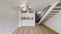 This two-floor residence offers a practical and functional layout spread across two levels. The main level centers around a generous carpeted living area illuminated by a ceiling fan with integrated lighting and a textured ceiling overhead. A pass-through kitchen window connects the kitchen to the living space, creating an open and communicative flow between the two areas. The kitchen is equipped with white appliances, including an electric coil-top range and an over-the-range Whirlpool microwave, complemented by warm oak-toned cabinetry offering ample storage. A dark wood entry door anchors the entryway near the base of the staircase, which ascends to the upper floor via carpeted steps and a sleek black metal railing. The upper level features additional carpeted space bathed in natural light from a large window dressed with vertical blinds, accompanied by another ceiling fan. A clean, white-tiled bathroom on the upper level includes a tub and shower combination with chrome fixtures, maintaining a bright and neutral aesthetic throughout. The overall design is clean and straightforward, with neutral tones, soft carpeting, and practical finishes that create a comfortable, move-in-ready living environment across both floors.