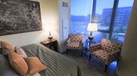 This thoughtfully designed space offers a harmonious blend of comfort and style, highlighted by expansive floor-to-ceiling windows that flood the room with natural light and frame striking views of the surrounding cityscape and greenery beyond. The bedroom area is anchored by a neatly made bed dressed in a textured grey coverlet with warm accent pillows, positioned alongside a classic wooden nightstand topped with a soft-glowing table lamp. A decorative city map serves as a focal point on the wall, adding a locally inspired, artistic touch to the space. Adjacent to the windows, a seating area features two matching upholstered armchairs in a bold geometric orange and white pattern, each accompanied by a coordinating side table and lamp — creating an inviting nook ideal for relaxation or conversation. The room is finished with a plush area rug that adds warmth and texture underfoot. The overall ambiance is cozy yet refined, balancing residential comfort with an elevated aesthetic. The generous window line ensures abundant natural light throughout the day while maintaining a strong visual connection to the urban environment outside.