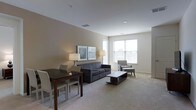 This single-floor space presents an open-plan layout that seamlessly integrates a living area with a dining space. The interior is finished with neutral beige walls and wall-to-wall carpeting, creating a warm, cohesive aesthetic throughout. Recessed lighting in the white ceiling supplements the natural light that enters through large, multi-pane windows positioned along one wall, providing a bright and airy atmosphere.

The living area is furnished with a dark grey sofa paired with a matching coffee table, while a light grey accent chair with an ottoman adds a secondary seating option. Two framed artworks and a pair of table lamps flank the sofa, adding visual interest and ambient lighting to the space.

Adjacent to the living area, a dark wood dining table with upholstered chairs defines the dining space, accommodating a small group comfortably. A laptop positioned on the table also suggests the space can double as a work area. Overall, the layout is efficiently arranged, making practical use of the available space while maintaining an uncluttered, modern feel.
