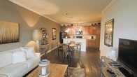 This single-floor residence showcases an open-concept floor plan that thoughtfully integrates the living, dining, and kitchen areas into one cohesive, flowing space. Rich, dark hardwood flooring extends throughout, providing a warm and grounded foundation that complements the neutral taupe wall tones and crisp crown molding accents.

The kitchen is a functional centerpiece, featuring warm honey-toned wood cabinetry that reaches toward the ceiling, offering generous storage. A peninsula with a light countertop serves as a natural divider between the kitchen and dining area, ideal for casual interaction. Black appliances, including a refrigerator and range, add a bold contrast, while pendant lighting above the peninsula provides focused task illumination.

The dining area is positioned just off the kitchen, accommodating a round table with metal-framed chairs beneath a stylish hanging light fixture — creating an inviting spot for everyday meals or entertaining.

The living area flows naturally from the dining space, furnished with a comfortable white sofa, layered area rugs, and eclectic decorative accents that add personality and depth to the room. The overall ambiance is warm and welcoming, with a curated blend of rustic and contemporary design elements that make this space feel both livable and stylish.