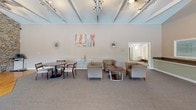 This single-floor community clubhouse offers a well-appointed gathering space designed for both comfort and functionality. The main lounge area is defined by soaring vaulted ceilings with exposed beam detailing and track lighting, creating a bright, airy atmosphere. A continuous bank of large windows along one wall frames sweeping views of the outdoor pool area, a gazebo, and surrounding greenery, filling the space with natural light throughout the day. Comfortable upholstered club chairs and low-profile tables are arranged throughout the lounge to encourage relaxed socializing, while a separate dining table and chair set provides additional seating options. The entryway features a striking glass-panel vestibule with dark metal framing, offering a clean, modern transition into the interior. Adjacent rooms are accessible via wide doorways, adding to the open flow of the space. The fully equipped kitchen is a standout feature, outfitted with warm maple cabinetry, dark countertops, hardwood-style flooring, and a full suite of black and stainless steel appliances including a refrigerator, dishwasher, microwave, and range. Crown molding adds a refined finishing touch to the kitchen area. Overall, the space presents a welcoming and versatile environment well suited for community events and gatherings.