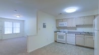 This single-floor residence offers a clean, functional layout designed for comfortable everyday living. The kitchen is a central feature of the space, outfitted with light wood cabinetry that lends a warm, understated aesthetic. White appliances — including a gas range, over-the-range microwave, and dishwasher — are neatly integrated beneath granite-toned countertops, providing a practical and cohesive workspace. The tile flooring in the kitchen is durable and easy to maintain, transitioning smoothly into carpeted areas beyond. A half-wall partition subtly defines the boundary between the kitchen and the adjacent living area, creating an open-concept feel while maintaining a sense of structure. The living space features neutral-toned carpet and continues the light, airy color palette established throughout. Bright white walls and flush ceiling lighting keep the interior feeling open and well-illuminated. Visible HVAC vents indicate climate control throughout the unit. An electrical panel access point is also conveniently located within the main space. Overall, the residence presents a straightforward, move-in-ready environment with a cohesive design that prioritizes practicality and ease of living.