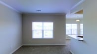 This single-floor home offers a welcoming and functional layout designed for comfortable everyday living. The interior is finished with warm, neutral-toned walls that create a calm and inviting atmosphere throughout. The living area is carpeted and bathed in natural light, courtesy of large windows that frame pleasant views of the surrounding neighborhood and lush greenery beyond. Crown molding accents the ceiling, adding a polished architectural detail that elevates the overall aesthetic. The space transitions smoothly into the kitchen area, which is visible through an open pass-through or peninsula, enhancing the sense of connectivity and openness between the rooms. The kitchen features light-colored countertops and cabinetry, complementing the neutral palette carried throughout the home. Additional windows in the kitchen area further brighten the space, ensuring that natural light reaches every corner. The flooring transitions from carpet in the living space to what appears to be a hard surface in the kitchen, offering both comfort and practicality. Ceiling vents are neatly integrated into the layout, supporting an efficient climate control system. Overall, this home presents a clean, move-in-ready environment with thoughtful design details and abundant natural light.