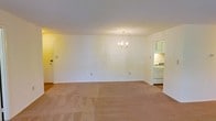 This single-floor residence offers a straightforward, functional layout centered around a spacious open living area. The room is finished with soft, neutral-toned wall-to-wall carpeting that lends a warm, understated feel to the space. White walls keep the interior bright and versatile, providing a clean backdrop adaptable to a variety of furnishing styles.

A classic candelabra-style chandelier serves as a focal point overhead, adding a subtle decorative touch to the otherwise minimalist room. A standard entry door is positioned along one wall, providing direct access to the living area.

An open passageway connects the living area to the kitchen, which is visible from the main room. The kitchen features white cabinetry, a white gas range, and light-colored walls, contributing to a cohesive, clean aesthetic throughout. The cabinetry provides ample storage, and the layout appears compact yet practical.

Overall, the space presents a simple, move-in-ready environment with a neutral palette and essential features, making it well-suited for those seeking a clean, functional living arrangement on a single level.