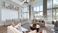 This elegantly designed single-floor space exudes a refined, contemporary aesthetic that immediately captures attention. The centerpiece of the lounge is its dramatically high ceilings, accentuated by recessed cove lighting that casts a warm, ambient glow throughout. Floor-to-ceiling windows and glass walls flood the interior with an abundance of natural light, offering sweeping views of lush palm trees and an outdoor area beyond, seamlessly blending the indoors with the surrounding environment.

The interior is thoughtfully appointed with a sophisticated neutral palette, featuring cream and taupe upholstered seating arrangements that include tiered lounge chairs and plush sofas adorned with decorative pillows. A striking organic wood and glass coffee table serves as a focal point, anchored by an artfully patterned area rug with flowing, marble-like swirls. Built-in wall shelving displays curated decorative objects, adding a layer of personalized elegance.

Sheer white drapery panels frame the expansive windows, softening the bright light while maintaining an open, airy feel. A ceiling-mounted projector hints at the space's versatility for presentations or entertainment. Wall-mounted accent lighting and thoughtful spatial planning round out this polished, multifunctional lounge, making it equally suited for relaxed gatherings and formal events.
