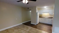 This single-floor apartment at Regency Apartments at Southland offers a practical, open layout that connects the main living area to the kitchen. The living space features light-toned tile flooring and is equipped with a ceiling fan with integrated lighting, providing both comfort and illumination. Neutral, light beige walls keep the space feeling bright and clean throughout.

The kitchen is accessible via an open passageway and is outfitted with white cabinetry offering ample storage above and below the countertops. A dark-toned countertop provides a subtle contrast against the white cabinetry, and a white refrigerator is positioned at the kitchen's entrance. The kitchen also includes a sink with visible hardware and under-cabinet lighting that adds a functional glow to the workspace.

The transition between the living area and the adjacent space is marked by a shift in flooring from tile to a darker hardwood-style surface, creating a clear visual distinction between zones. White baseboards run throughout, adding a clean, finished look to the overall interior. The layout is straightforward and well-suited for everyday living.