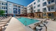 This expansive outdoor courtyard amenity space in Salt Lake City, Utah, is designed to deliver a resort-style experience within a contemporary multi-story residential community. At its heart lies a generously sized swimming pool with crystal-clear blue water, complemented by a separate spa that features elegant stone tile surrounds. The pool deck is lined with rows of cushioned chaise lounges, providing ample space for sunbathing and relaxation beneath the open Utah sky.

Flanking the pool area, wooden pergola structures offer shaded gathering spots, adding warmth and architectural character to the hardscaped surroundings. A dedicated lounge zone features comfortable outdoor seating arranged around a sleek rectangular fire pit, creating an inviting atmosphere for evening socializing. Mature trees and low-water landscaping with decorative gravel accents introduce natural elements that soften the urban setting.

The courtyard is framed on all sides by the multi-story building, whose façade blends blue paneling, white lap siding, and warm sandy brick — a modern design palette that gives the community a distinctive, contemporary aesthetic. Private balconies overlook the courtyard from multiple levels, enhancing the sense of connectivity between indoor and outdoor living. The layout is thoughtfully designed to foster community while offering quiet corners for individual retreat.