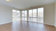 This 2-bedroom, 2-bathroom residence in Bedford is designed to maximize natural light and openness. The living area is a standout feature, showcasing a wraparound arrangement of large, floor-to-ceiling windows fitted with horizontal blinds, flooding the space with daylight and offering views of the surrounding Bedford neighborhood, including glimpses of nearby buildings and greenery. The corner window configuration creates a bright, airy atmosphere that enhances the sense of spaciousness throughout the room.

The flooring consists of smooth, light-toned hardwood-style planks that run uniformly across the living area, contributing to a clean and contemporary aesthetic. The neutral wall tones and white ceiling complement the minimalist design, creating a versatile backdrop suitable for a variety of furnishing styles.

Ceiling-mounted HVAC vents are integrated discreetly into the layout, ensuring functional climate control without disrupting the clean lines of the interior. The overall ambiance of the space is modern and understated, with an emphasis on natural light, open sightlines, and a polished, move-in-ready finish throughout.