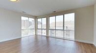This 2-bedroom, 2-bathroom residence in Bedford is designed to maximize natural light and openness. The living area is a standout feature, showcasing a wraparound arrangement of large, floor-to-ceiling windows fitted with horizontal blinds, flooding the space with daylight and offering views of the surrounding Bedford neighborhood, including glimpses of nearby buildings and greenery. The corner window configuration creates a bright, airy atmosphere that enhances the sense of spaciousness throughout the room.

The flooring consists of smooth, light-toned hardwood-style planks that run uniformly across the living area, contributing to a clean and contemporary aesthetic. The neutral wall tones and white ceiling complement the minimalist design, creating a versatile backdrop suitable for a variety of furnishing styles.

Ceiling-mounted HVAC vents are integrated discreetly into the layout, ensuring functional climate control without disrupting the clean lines of the interior. The overall ambiance of the space is modern and understated, with an emphasis on natural light, open sightlines, and a polished, move-in-ready finish throughout.