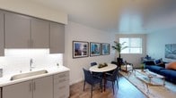 This single-floor unit showcases a cohesive modern design with light wood-style flooring, neutral walls, and clean lines carried throughout every room. The kitchen is well-appointed with floor-to-ceiling grey cabinetry, stainless steel appliances including a refrigerator, electric range, microwave, and dishwasher, paired with white quartz countertops and a classic white subway tile backsplash with under-cabinet lighting. A deep single-basin sink completes the functional layout.

Adjacent to the kitchen, the dining area accommodates a round white table with seating for four, while framed artwork adds a curated touch to the walls. The living area receives natural light through a large window and offers comfortable space for seating arrangements alongside additional wall art and greenery.

The bedroom features dark wood-style flooring, a generous closet with sliding doors, and softly lit wall space ideal for personal decor. The bathroom is finished in crisp white tones with a soaking tub and shower combination, a curved curtain rod, grey vanity cabinetry, white countertops, and a wide mirror, creating a bright, clean, and functional space.