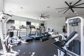 Located on Cole Drive in Barnegat, NJ, this single-story fitness center offers a comprehensive workout environment designed for both cardio and strength training. The space is finished with light blue-gray walls and durable rubber flooring throughout, creating a clean, functional atmosphere that is both practical and visually cohesive.

The main workout area is generously proportioned, accommodating a full array of equipment including multiple treadmills, elliptical machines, and a variety of strength-training stations such as cable machines, leg press equipment, and free weight racks with dumbbells. Large multi-pane windows line the walls, flooding the space with abundant natural light and offering views of the surrounding greenery, contributing to an open and inviting atmosphere.

Overhead, large-blade ceiling fans are strategically positioned to promote airflow across the workout floor, complemented by recessed lighting for consistent illumination. A mirrored wall in one section expands the visual depth of the room. A pass-through window connects to an adjacent room, and a clearly marked exit door ensures safe accessibility. The layout is thoughtfully arranged to provide ample circulation space between equipment, supporting a comfortable and efficient fitness experience.