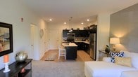 This single-floor space showcases an open-concept layout that seamlessly connects the living area to the kitchen, creating a functional and cohesive environment. The kitchen is a standout feature, equipped with dark, floor-to-ceiling cabinetry that provides a striking contrast against the light-toned countertops and white walls. Stainless steel appliances, including a refrigerator, microwave, and range, lend a modern, polished aesthetic. A central island with a breakfast bar offers additional prep space and casual seating for two, illuminated by pendant lighting overhead. Recessed ceiling lights are distributed throughout, ensuring consistent, well-distributed illumination across the space. The flooring transitions from light hardwood in the kitchen and entryway to plush carpet in the living area, defining each zone while maintaining visual continuity. The entryway, visible from the main living space, features white paneled doors and provides a clean, welcoming arrival point. The open layout and neutral color palette give the space a bright, airy feel, making it feel open and easy to navigate. Overall, the design balances practicality with a contemporary aesthetic.