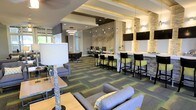 This single-floor space is a well-appointed communal lounge designed with a modern and sophisticated aesthetic. The open layout seamlessly integrates multiple functional zones, creating a versatile environment suited for both relaxation and productivity. Exposed beam detailing on the ceiling adds architectural interest, while layered lighting — including pendant fixtures, recessed lights, and wall sconces — contributes to a warm, inviting atmosphere throughout.

A central seating area features plush grey sofas and armchairs arranged around a low coffee table, anchored by a bold geometric-patterned carpet in grey and green tones that defines the space. Adjacent to this lounge area is a bar-height counter lined with black and green upholstered stools, accented by stacked stone columns and wall-mounted flat-screen televisions, lending a hospitality-inspired feel.

A dedicated workstation zone is positioned toward the rear, offering individual desk spaces with chairs, making it ideal for focused tasks. A sculptural wall accent adds an artistic focal point, while natural light filters in through windows, brightening the overall space. The combination of textures — stone, fabric, and wood — creates a layered, contemporary environment.