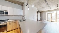 Located at 2200 Market in Denver's vibrant Curtis Park neighborhood, this one-bedroom, one-bathroom unit — known as the Mineral floor plan — showcases a modern industrial aesthetic throughout. Exposed concrete ceilings with visible ductwork and conduit lend an authentic loft character, while warm hardwood flooring grounds the space with a welcoming contrast.

The open-concept kitchen is a standout feature, anchored by a large quartz-topped island with bar seating, a stainless steel undermount sink with a matte black faucet, and a white subway tile backsplash. Natural wood cabinetry is paired with sleek white upper cabinets, and a full suite of stainless steel appliances — including a French door refrigerator, gas range, and over-the-range microwave — complete the well-appointed space. Glass pendant lights hang above the island, adding an industrial-chic finishing touch.

The living area is bright and airy, bathed in natural light from large windows and a set of French doors that open to views of the Denver skyline. Track lighting and a concrete ceiling continue the industrial theme seamlessly. A unique built-in ladder leads to elevated storage shelving in the closet area, maximizing vertical space creatively.

The bathroom features a combination tub and shower with matte black fixtures, clean white walls, and hardwood-style flooring. A spacious walk-in closet with wire shelving provides ample storage.
