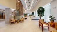 Located at 19 NE First Ct in Dania Beach, Florida, this sophisticated single-floor lobby space offers a striking first impression through its contemporary design and carefully curated interiors. The space is defined by soaring double-height ceilings that create an airy, open atmosphere throughout. Large-format neutral tile flooring extends across the entire area, providing a clean, seamless foundation that enhances the sense of expansiveness.

A dramatic floor-to-ceiling wood lattice feature wall serves as the architectural centerpiece, adding warmth and texture to the otherwise light and bright palette. Suspended circular ring chandeliers cascade from the ceiling, providing both ambient illumination and a bold design statement. Multiple distinct seating zones are thoughtfully arranged throughout the space, including plush white sofas, a sage green banquette, cognac leather seating, and natural wood-framed accent chairs, offering a variety of comfortable gathering areas.

Floor-to-ceiling windows along the perimeter flood the interior with natural light and frame views of the outdoors. Lush indoor trees and tropical greenery are strategically placed to bring nature inside. A wood-paneled accent wall with a large display screen serves as a functional focal point near the entrance. The overall ambiance is refined yet welcoming, blending South Florida's tropical character with upscale, modern hospitality design.