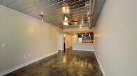 This single-floor space blends industrial character with modern functionality, creating a distinctive and open atmosphere throughout. The living area commands attention with its polished concrete floors, which reflect light and lend a sleek, urban feel to the interior. Overhead, a corrugated metal ceiling adds a bold industrial aesthetic, complemented by exposed round ductwork that runs the length of the space. A ceiling fan with integrated lighting anchors the main living area, while a rail track lighting system provides flexible, directional illumination along the ceiling. The open layout flows seamlessly toward the kitchen, which is defined by a white breakfast bar counter that serves as a natural dividing element between the living and culinary spaces. Dark cabinetry lines the kitchen walls, offering generous storage, while pendant lighting descends above the bar area, adding warmth and task-focused light. Recessed ceiling lights in the kitchen zone ensure the workspace is well-lit. A white refrigerator and integrated appliances complete the kitchen's functional design. Toward the rear of the space, doorways lead to additional rooms, including what appears to be a bathroom and further private areas. Neutral-toned walls throughout keep the overall ambiance bright and adaptable, allowing the industrial architectural details to remain the defining feature of this well-designed space.