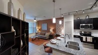 Located in Carrollton, this single-floor residence offers a thoughtfully designed open-concept layout that integrates the kitchen, dining, and living spaces into one cohesive and inviting environment. The kitchen is appointed with sleek granite countertops, dark cabinetry, a stainless steel sink, and pendant lighting that adds a contemporary touch. A breakfast bar with stylish seating provides a casual dining option and serves as a natural transition point between the kitchen and living area. The living room exudes warmth through a rich accent wall in a warm earth tone, complemented by wood-look flooring and a plush area rug. A ceiling fan and large windows fitted with blinds allow for both comfort and natural light throughout the day. Decorative elements, including patterned wall panels and curated artwork, add visual interest and personality to the space. Track lighting runs along the ceiling, ensuring the interiors are well-lit and functional. The overall ambiance is modern yet welcoming, balancing stylish design choices with everyday livability. This Carrollton residence is well-suited for those seeking a contemporary, open, and connected living experience.
