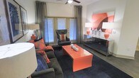 This single-floor space makes a striking first impression with its bold, contemporary interior design. The living area is anchored by a rich charcoal sectional sofa adorned with vibrant orange and patterned throw pillows, creating a dynamic contrast that defines the room's energetic color palette. A glossy orange coffee table serves as the focal centerpiece, complementing the coordinated décor throughout.

The space features hardwood flooring topped with a deep charcoal area rug, adding warmth and texture underfoot. A ceiling fan with an integrated light fixture provides both comfort and practical illumination. Natural light pours in through multiple large windows dressed with floor-length gray drapery panels, lending an airy, open feel to the interior.

A sleek mirrored console table lines one wall, styled with decorative lamps, ceramic accents, and a bold orange wall art piece that ties the room's color story together cohesively. A classic dark leather accent chair with an orange cushion detail rounds out the seating arrangement, positioned near the windows to take advantage of the natural light. The overall ambiance is modern and sophisticated, balancing bold pops of color with neutral tones to create a space that is both visually engaging and inviting.