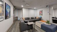 Located at 900 Carnarvon St in New Westminster, BC, this single-floor communal lounge offers a thoughtfully designed space for relaxation and social gathering. The layout is divided into two distinct seating zones, each with its own character and purpose.

The first zone features a large, deep-seated sectional sofa in charcoal grey, accented with warm tan throw pillows and complemented by a sleek low-profile side table. Colorful modular ottomans in blue and orange add a vibrant touch, while a laptop station nearby provides a functional element to the comfortable setting.

The second zone transitions into a more intimate arrangement of black leather armchairs with grey upholstered headrests, organized in a semi-theater configuration. Colorful ottomans in yellow and orange punctuate the seating, and a small side table adds practicality. A potted plant introduces a natural element to the contemporary interior.

Throughout the space, framed classic movie posters — including Casablanca, Star Wars, E.T., Top Gun, and The Shawshank Redemption — lend a cinematic theme and visual interest to the neutral walls. The dropped tile ceiling features recessed lighting and integrated HVAC, while the patterned carpet with yellow-green accents grounds the modern aesthetic. Slim vertical windows along one wall allow soft natural light to filter in, enhancing the welcoming ambiance of this versatile lounge.