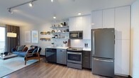 This well-appointed 1-bedroom, 1-bathroom residence offers a thoughtfully designed living space with a clean, modern aesthetic. The open-concept kitchen serves as a focal point, outfitted with stainless steel appliances including a full-size refrigerator, electric range with oven, built-in microwave, and dishwasher. Crisp white upper cabinetry extends to the ceiling, maximizing storage, while sleek dark lower cabinets provide striking contrast. Open floating shelves mounted on the wall add a contemporary touch, offering both functional storage and display space. Light-colored countertops and a white subway tile backsplash contribute to the kitchen's bright, cohesive look. Recessed lighting and track lighting illuminate the space efficiently. Smooth light wood-tone flooring runs throughout, adding warmth to the modern palette. The living area is visible from the kitchen, reinforcing the open and connected feel of the floor plan. The overall ambiance is clean, urban, and functional — ideal for those who appreciate contemporary design paired with practical, well-organized living spaces.