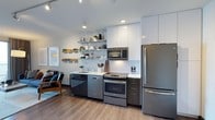 This well-appointed 1-bedroom, 1-bathroom residence offers a thoughtfully designed living space with a clean, modern aesthetic. The open-concept kitchen serves as a focal point, outfitted with stainless steel appliances including a full-size refrigerator, electric range with oven, built-in microwave, and dishwasher. Crisp white upper cabinetry extends to the ceiling, maximizing storage, while sleek dark lower cabinets provide striking contrast. Open floating shelves mounted on the wall add a contemporary touch, offering both functional storage and display space. Light-colored countertops and a white subway tile backsplash contribute to the kitchen's bright, cohesive look. Recessed lighting and track lighting illuminate the space efficiently. Smooth light wood-tone flooring runs throughout, adding warmth to the modern palette. The living area is visible from the kitchen, reinforcing the open and connected feel of the floor plan. The overall ambiance is clean, urban, and functional — ideal for those who appreciate contemporary design paired with practical, well-organized living spaces.