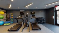 Located at 27W555 High Lake Rd in Winfield, IL, this single-floor fitness center is a well-appointed facility designed to support a comprehensive workout experience. The space features a contemporary aesthetic with dark charcoal-toned walls, recessed lighting, and linear LED fixtures that create a focused, energetic atmosphere throughout.

The cardio zone is equipped with multiple treadmills, an elliptical cross-trainer, and upright stationary bikes, all positioned to take advantage of floor-to-ceiling glass windows that overlook an outdoor pool and patio area, providing ample natural light and scenic views. Wall-mounted flat-screen televisions are positioned above the cardio machines for entertainment during workouts.

A dedicated strength training area includes a multi-function cable machine and a well-stocked rack of dumbbells, offering a range of resistance options for users of all fitness levels. Full-length mirrors line one wall, enhancing the sense of space and allowing for proper form checks during exercise.

A separate functional training zone, enclosed by sleek black-framed glass partitions, houses stability balls, resistance accessories, and open floor space ideal for stretching, yoga, or group exercise. The durable, textured flooring throughout the facility provides both comfort and safety. Together, these areas form a cohesive, modern fitness environment suited for diverse training needs.