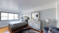 This single-floor space showcases a well-appointed bedroom designed with a refined, glamorous aesthetic. The room is anchored by a tufted upholstered bed with a classic camelback headboard finished in silver fabric, complemented by layered decorative pillows in coordinating grey and white tones. A large, mirrored dresser with multiple drawers serves as a functional yet decorative focal point along the main wall, topped with a silver-framed mirror that reflects light throughout the space. The warm hardwood flooring provides a rich contrast to the cool silver and grey color palette of the furnishings, while a plush dark shag area rug adds texture and depth underfoot. Walls are painted in a soft, neutral grey-white tone, contributing to the room's calm and sophisticated ambiance. A window fitted with wood-slat blinds allows for adjustable natural light. Ample storage is provided by the multi-drawer dresser, and additional seating is suggested by a velvet accent chair visible at the edge of the frame. Overall, the bedroom presents a cohesive, upscale interior with attention to coordinated finishes and comfortable furnishings.