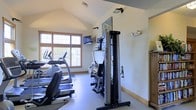 This dedicated fitness space is thoughtfully designed to support a comprehensive workout routine. The room features vaulted ceilings that create an open, airy atmosphere, enhancing the sense of space and making exercise sessions feel less confined. Large wood-framed windows along one wall allow generous natural light to fill the room, offering views of the surrounding greenery and creating an inviting environment for physical activity.

The space is outfitted with a selection of cardio equipment, including a treadmill and an elliptical trainer, positioned to take advantage of the natural light and outdoor views. A multi-functional cable resistance machine serves as a centerpiece for strength training, offering versatile options for a full-body workout. A wall-mounted flat-screen television provides entertainment during exercise sessions, while wall-mounted equipment hooks keep accessories organized and within reach.

The flooring is finished with a smooth, durable surface suited to fitness use, and the neutral, light-toned walls contribute to a clean and focused atmosphere. Subtle track lighting supplements the natural light as needed. The overall design strikes a balance between functionality and comfort, making this fitness room a practical and motivating space for daily exercise.